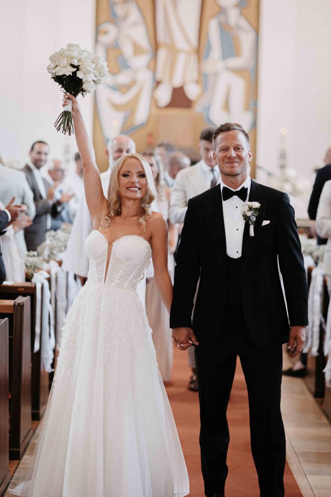 Beim Auszug aus der Kirche geht das Brautpaar nebeneinander Richtung Ausgang. Die Braut hält den Strauß mit dem gestreckten Arm in die Luft.